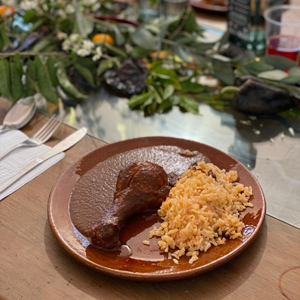 mole sauce over a chicken drumstick next to a side of orange rice on a brown plate sitting on a wooden table with silverware and greenery in the background