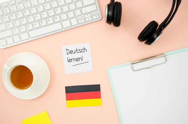 Deutsch lernen! on a post-it note, keyboard, headphones, a cup of tea, a small German flag, and a clipboard